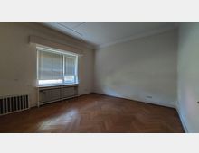 Leerer Raum mit weiß gestrichenen Wänden, einem großen Fenster mit heruntergelassenen Jalousien und einem Heizkörper darunter. Der Boden besteht aus Parkett in Fischgrätmuster, die Beleuchtung ist minimal, und es gibt einige Steckdosen an den Wänden.