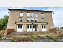 Zweistöckiges Gebäude mit einer schlichten Fassade, mehreren großen Fenstern und weißen Eingangstüren im Erdgeschoss. Der Zugang erfolgt über eine breite, mit Ziegelsteinen gepflasterte Treppe, die von niedrigen Ziegelmauern flankiert wird; im Hintergrund sind Bäume und Himmel erkennbar.