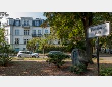 Ein kleiner Platz mit Bäumen und einem Stein mit Reliefskulptur im Vordergrund, daneben ein Schild mit der Aufschrift „Pestalozziplatz“. Im Hintergrund steht ein historisches, weißes Gebäude mit dekorativen Fassadenelementen und Balkonen.
