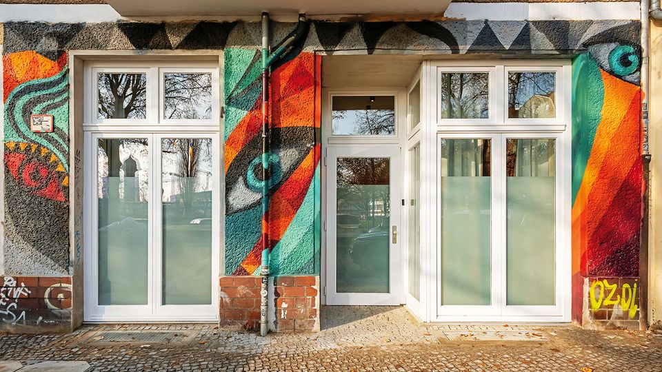 Eingangsbereich eines Wohn- oder Geschäftsgebäudes mit weiß gerahmter Glastür und großen Fenstern, umgeben von einer bunt bemalten Graffiti-Fassade. Vor dem Eingang liegt ein gepflasterter Gehweg, in den Scheiben spiegeln sich Bäume und die Straße.
