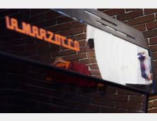 Nahaufnahme eines Spiegels mit der reflektierten Schrift "La Marzocco" in orange. Im Hintergrund ist eine Backsteinwand sowie Kleidung auf einem Kleiderbügel zu sehen.