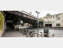 Straßenszene vor dem S-Bahnhof Yorckstraße in Berlin, mit einer Brücke aus dunklem Stahl über der Straße, Hinweisschildern für die S-Bahn und einem Gebäude mit auffälliger Wandbeschriftung. Fahrräder parken entlang des Bürgersteigs, eine Person ist auf dem Gehweg unterwegs.