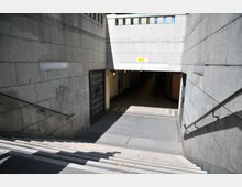 Treppenabgang zu einer Unterführung mit grauen Steinwänden, Handläufen an beiden Seiten und einem schattigen Eingang im Hintergrund. Links und rechts der Unterführung befinden sich Gittertüren, während darüber ein Geländer sichtbar ist.