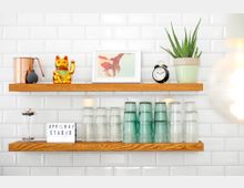 Zwei hölzerne Wandregale vor einer weißen gekachelten Wand. Auf dem oberen Regal stehen ein Kupferkrug, eine goldene Winkekatze, ein gerahmtes Foto, eine kleine schwarze Uhr und ein grüner Blumentopf mit einer Pflanze. Auf dem unteren Regal stehen gestapelte Glasbecher in klaren und grünen Tönen sowie ein kleines Schild mit der Aufschrift „APRILMAY STUDIO“.