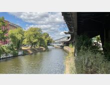 Ein Fluss in einer urbanen Umgebung, gesäumt von Bäumen und einem rosafarbenen Gebäude auf der linken Seite. Rechts verläuft eine erhöhte Bahntrasse, die teilweise einen modernen Brückenbau über den Fluss einschließt.