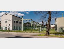 Außenansicht einer Umspannstation mit metallischen Stromleitungen und grünen Stützpfeilern, umgeben von einem Zaun. Im Vordergrund steht ein Baum, im Hintergrund sind ein weißes Gebäude und weitere elektrische Anlagen sichtbar.