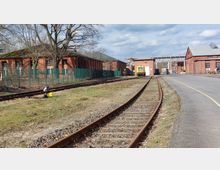 Bahngelände mit Gleisen, die an mehreren Backstein-Gebäuden entlangführen; im Hintergrund steht ein gelbes Schienenfahrzeug vor einer Halle. Ein Zaun trennt die Gebäude auf der linken Seite von den Gleisen, während rechts eine asphaltierte Straße verläuft.