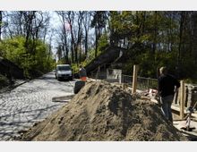 Baustelle an einer Kopfsteinpflasterstraße in einem bewaldeten Bereich, mit einem Sandhaufen im Vordergrund, Bauarbeitern und Baumaterialien. Im Hintergrund führen Treppen mit Graffiti beschmierten Mauern einen Hügel hinauf, und ein weißer Lieferwagen steht auf der Straße.