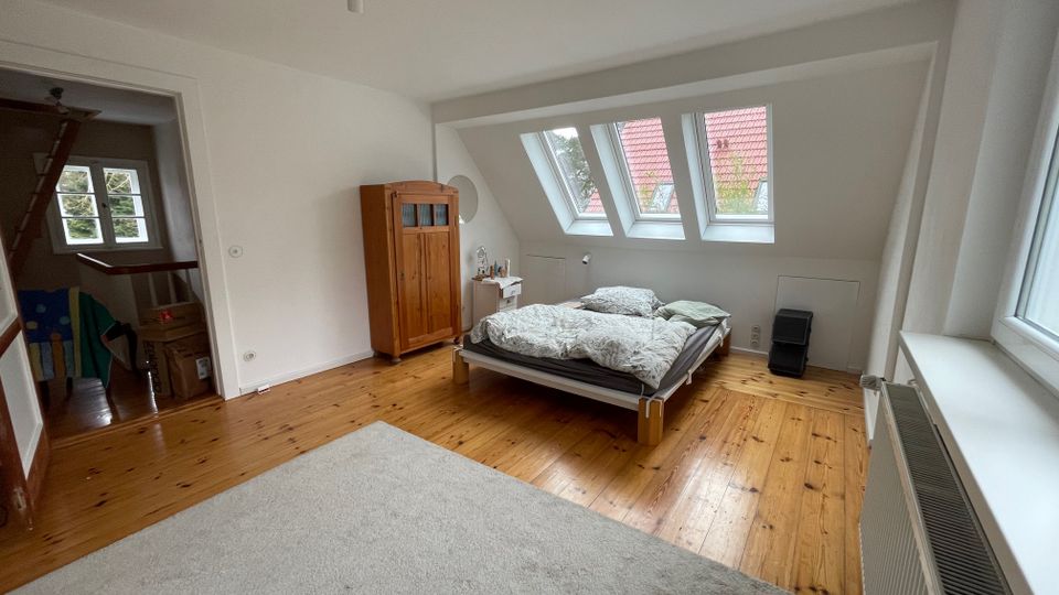 Helles Dachgeschoss-Schlafzimmer mit Holzfußboden, niedrigem Bett und Kleiderschrank. An der schrägen Dachwand befinden sich mehrere Dachfenster, daneben ein großes Seitenfenster und ein offener Durchgang zu einem angrenzenden Raum.