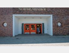 Eingang des Nordbahnhofs in Berlin, mit markanten orangefarbenen Türen und einem Klinkerfassaden-Design. Über dem Eingang befindet sich der Schriftzug "NORDBAHNHOF" in goldenen Buchstaben, umgeben von runden Fensterelementen in den Ziegelwänden.