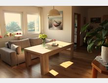 Helles Wohnzimmer mit einem großen Holztisch in der Mitte, einer grauen Eckcouch und einem Bild an der Wand, das ein Boot zeigt. Große Fenster lassen Tageslicht herein, und im Raum stehen mehrere Zimmerpflanzen sowie dekorative Elemente.