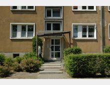 Eingang eines Wohnhauses mit gelblicher Putzfassade, weißen Fenstern mit Gardinen und einem kleinen Vordach aus Metall und Glas. Vor dem Gebäude befinden sich Treppenstufen, ein gepflasterter Weg und gepflegte Büsche sowie Blumenbeete.