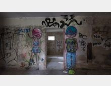 Ein verlassenes Gebäude mit bemalten, verfallenen Wänden, auf denen Graffiti und bunte Wandmalereien von zwei stilisierten Figuren zu sehen sind: links ein Mädchen mit rosa Haaren und rechts ein Junge mit blauen Haaren. Im Hintergrund ein Durchgang mit einem kleinen Fenster, das etwas Licht in den Raum wirft.