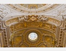 Innenansicht einer prunkvollen Kirchenkuppel mit kunstvollen Verzierungen aus vergoldetem Stuck, Engelfiguren und detaillierten Reliefs. In der Mitte befindet sich ein rundes Buntglasfenster, das Tageslicht hereinlässt.