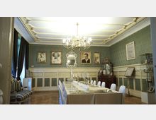 Eleganter Raum mit grüner Tapete und weißen Zierleisten, einer langen, eingedeckten Tafel mit Kerzenhaltern und einem Kronleuchter an der Decke. Wände sind mit gerahmten Porträts, einem Spiegel und historischen Ausstellungstafeln dekoriert.