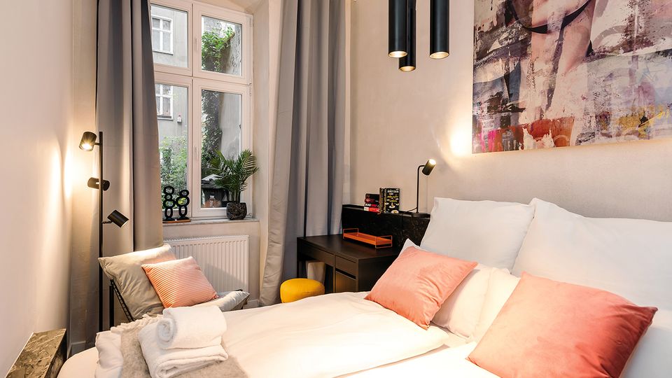 Modernes Schlafzimmer mit Doppelbett, weißen Bettdecken und rosafarbenen Kissen, dazu graue Vorhänge und ein großes Fenster mit Blick in einen begrünten Innenhof. Im Raum stehen ein schwarzer Schreibtisch mit Hocker, mehrere schwarze Lampen und ein großes Wandbild über dem Bett.