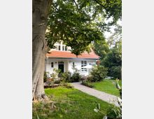 Ein weißes Einfamilienhaus mit rotem Ziegeldach, umgeben von einem Garten mit Rasen, kleinen Sträuchern und einem großen Baum im Vordergrund. Ein gepflasterter Weg führt zur Eingangstür, daneben stehen Gartenmöbel und ein Sonnenschirm.