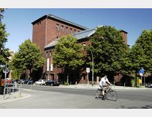 Backsteingebäude mit Flachdach und großen Fenstern, umgeben von Bäumen an einer Straßenecke; im Vordergrund fahren ein Radfahrer und parkende Autos.