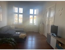 Wohnzimmer mit großen Fenstern, durch die viel Tageslicht einfällt. Im Raum stehen ein dunkles Ecksofa, ein flacher Fernseher auf einem Holz-TV-Schrank, weiße Türen mit Glaseinsätzen sowie ein Bücherregal; Holzboden und dekorative Elemente wie Pflanzen und Wandkunst sind sichtbar.