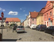 Straßenszene in einer historischen Altstadt mit gepflastertem Boden, geparkten Autos und bunten Gebäuden mit verzierten Fassaden in Gelb, Pink und Rot. Im Hintergrund sind ein Gebäude mit rotem Satteldach und ein Straßencafé mit Sonnenschirmen zu sehen, auf dem blauen Himmel ziehen vereinzelt Wolken.