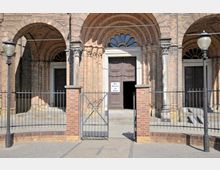 Historische Kirchenfassade aus Ziegelstein mit Rundbögen, dekorativen Säulen und einem Holzeingangstor, auf dem ein Schild mit der Aufschrift „Die Kirche ist offen“ angebracht ist. Im Vordergrund steht ein schmiedeeisernes Tor mit niedriger Backsteinmauer und zwei Laternenpfählen.