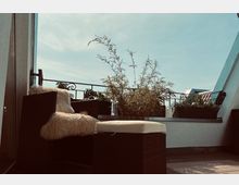 Dachterrasse mit einem geflochtenen Sessel, der mit einem Fell dekoriert ist, sowie einem kleinen Hocker mit Sitzpolster. Im Hintergrund sind Pflanzen in Blumenkästen, ein schmiedeeisernes Geländer und ein heller Himmel zu sehen.
