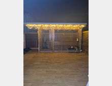 Holzhütte mit Glas- und Holzverkleidung, beleuchtet durch eine Lichterkette am Dachrand. Der Vorderbereich besteht aus einer Holzbodenfläche, daneben befinden sich kleine Möbelstücke und Dekorationen.