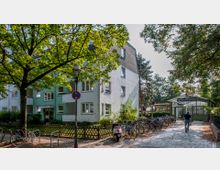 Wohngebäude mit heller Fassade und Eckbalkonen auf der linken Seite, daneben der Eingang des S-Bahnhofs Berlin-Lichterfelde West mit einem gläsernen Vordach. Im Vordergrund sind ein Baum, geparkte Fahrräder und ein Fußgänger auf einem gepflasterten Weg zu sehen.