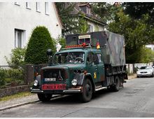Ein historischer, dunkelgrüner Lkw mit Tarnmuster-Aufbau steht am Straßenrand einer Wohngegend. Im Hintergrund sind weiße Häuser mit roten Dächern, dichtes Grün und geparkte Fahrzeuge zu sehen.