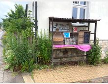 Holzüberdachte Verkaufshütte mit dekorativen Schildern und Kleingegenständen, darunter ein handgeschriebenes Menü auf einer Tafel. Die Hütte grenzt an ein Wohnhaus mit weißer Putzfassade, umgeben von üppigem Grün und Wildpflanzen neben einem Gehweg mit Pflastersteinen.