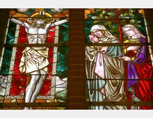Buntglasfenster mit christlicher Szene: Links ist die Kreuzigung Jesu mit Dornenkrone und Lendentuch dargestellt, rechts stehen zwei Frauen in andächtiger Haltung, eine mit gefalteten Händen. Der Hintergrund zeigt farbige Muster mit Blatt- und geometrischen Motiven.