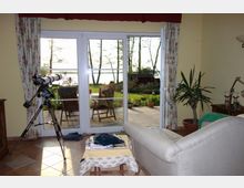 Wohnzimmer mit hellen Wänden und Fliesenboden, das durch eine große Glasschiebetür auf eine Terrasse mit Gartenmöbeln und Blick auf einen See führt. Im Raum befinden sich ein Sofa, ein kleiner Tisch, ein Teleskop und eine große Zimmerpflanze.