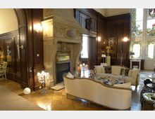 Eleganter Wohnraum mit holzvertäfelten Wänden, einem großen steinernen Kamin und dekorativen Glasfenstern. Zentral stehen beige Sofas um einen Couchtisch, umgeben von Kerzenhaltern und klassischer Beleuchtung.