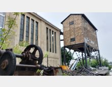 Industriegelände mit einem historischen Fabrikgebäude und einem erhöhten Holzkonstrukt auf Metallstützen. Im Vordergrund sind alte Maschinenteile und Schrott zu sehen, umgeben von vereinzeltem Grün.