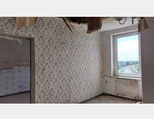 Ein heruntergekommenes Zimmer mit abblätternder Tapete im floralen Muster, einem beschädigten Deckenbereich, und einem Fenster mit Blick auf eine ländliche Straße und Landschaft. Links ist eine offene Tür zu einem weiteren Raum sichtbar.