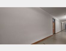 Langer, schlicht gestalteter Innenflur mit weiß gestrichenen Wänden und beigen Böden. Rechts befinden sich Holztüren mit Glaseinsätzen, die gleichmäßig entlang der Wand angeordnet sind.