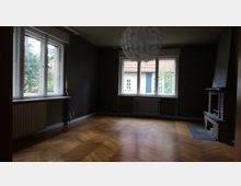 Ein leerer Wohnraum mit dunklen Wänden, großen Fenstern, die viel Tageslicht einlassen, und Blick auf ein Nachbarhaus mit roten Ziegeldächern. Der Raum verfügt über einen Kamin aus Ziegelsteinen auf der rechten Seite, einen Parkettboden und eine große Deckenleuchte.