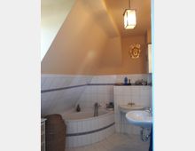 Kleines Badezimmer mit Dachschräge, einer Eckbadewanne mit weißen Fliesen und blauem Dekorband, einem Waschbecken und einer hängenden Deckenleuchte. Im Hintergrund ist eine goldfarbene Wanddekoration sichtbar.