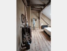 Schlafzimmer in einem Dachgeschoss mit schrägen Decken und sichtbaren Holzträgern. Der Raum hat einen Holzboden, eine schlichte Einrichtung mit einem Bett, einem kleinen Kleiderständer, einem dunklen Sideboard und einer Vase mit getrockneten Zweigen.