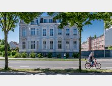 Straßenszene mit einer historischen Fassadenfront eines hellblauen, verzierten Wohngebäudes im Stil des 19. Jahrhunderts; im Vordergrund ein Fahrradfahrer auf einer Straße und eine Fußgängerin auf dem Gehweg, umgeben von Bäumen. Rechts daneben ist ein langes, rotes Backsteingebäude sichtbar.