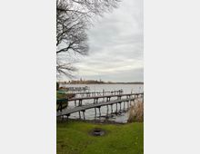 Blick auf einen See mit mehreren schmalen Holzstegen, die ins Wasser führen. Im Hintergrund sind am Ufer Bäume, Gebäude und Kirchtürme zu sehen; kahles Geäst eines Baumes ragt von der Seite ins Bild.
