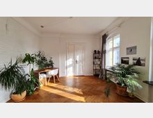 Ein heller Wohnraum mit weißen Wänden und Fischgrät-Parkettboden. Der Raum ist mit großen Fenstern, einem weißen Doppeltür-Eingang, Pflanzen in Töpfen, einem Holztisch mit Stuhl und einem schwarzen Regal mit Büchern und Dekoration ausgestattet.