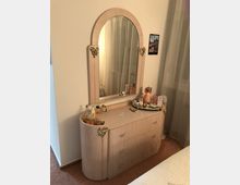 Ein beiger Frisiertisch mit integriertem ovalem Spiegel und goldfarbenen Verzierungselementen, auf dem verschiedene Parfümflaschen, Schmuck und Dekorationsgegenstände stehen. Der Tisch steht in einem Schlafzimmer mit cremefarbenen Wänden und bodenlangen Vorhängen.