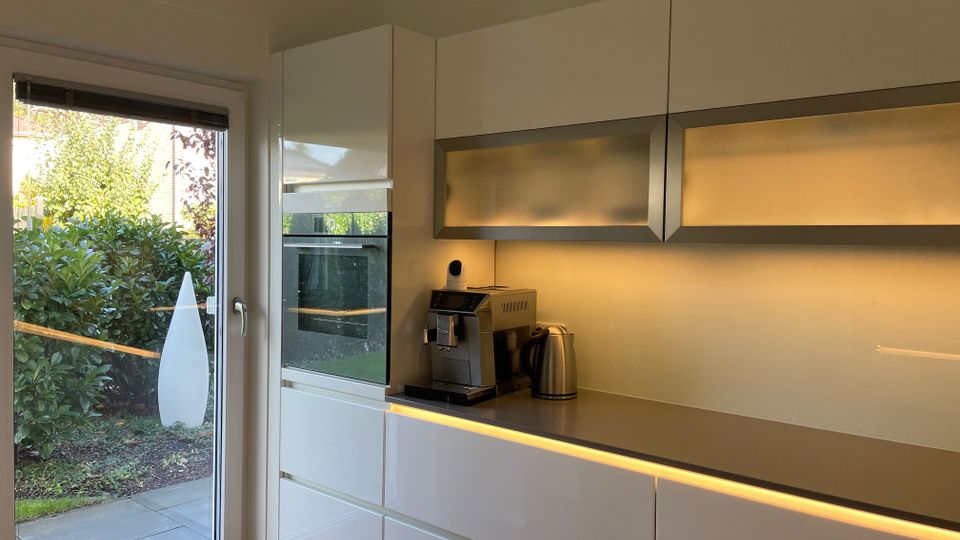 Moderne Küche mit weißen Hochglanzschränken und eingebautem Backofen. Eine Glastür führt zu einem Garten mit dichtem Grün, während warmes Unterbau-Licht die Arbeitsflächen und Küchengeräte beleuchtet.