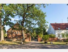 Eine historische Hofanlage mit einem roten Backsteingebäude links und einem weißen Putzbau mit rotem Ziegeldach rechts, umgeben von großen Bäumen und einer gepflasterten Zufahrt. Im Hintergrund ist ein offenstehendes Tor in einer Backsteinmauer zu sehen.