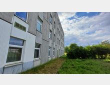 Mehrstöckiges Gebäude mit grauer, schlichter Fassade und rechteckigen Fenstern, umgeben von einer Grünfläche mit Gras und Sträuchern. Der Himmel ist bewölkt mit vereinzelten blauen Bereichen.