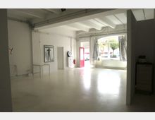 Ein heller, leerer Raum mit weiß gestrichenen Wänden und einer glatten, glänzenden Bodenoberfläche. Große Fenster mit Blick nach draußen, ein roter Türrahmen, Heizkörper an den Wänden und sichtbare Rohrleitungen sowie ein gerahmtes Poster und dekorative Elemente.