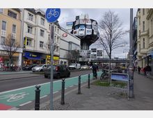 Straßenszene in einer urbanen Umgebung mit mehrstöckigen Gebäuden, Geschäften (u. a. Tchibo und C&A), einem markanten Hochhaus mit futuristischer Architektur im Hintergrund und einem Fahrradweg im Vordergrund. Auf der Straße sind Autos und Fahrräder zu sehen, während Gehsteige mit Fahrradbügeln und Fußgängern belebt sind.