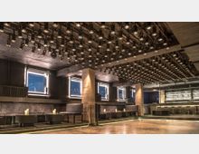 Ein moderner Nachtclub mit industriellem Design, gekennzeichnet durch Betonpfeiler, eine Vielzahl von Strahlern an der Decke und minimalistische Sitzbereiche mit Kerzen. Im Hintergrund befindet sich eine beleuchtete Bar mit hoch aufgereihten Getränkeflaschen und Barhockern.