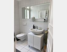 Helles, weiß gefliestes Badezimmer mit wandhängendem WC und Waschtisch auf einem Unterschrank. Über dem Waschbecken befindet sich ein breiter Spiegel mit Glasablage; im Hintergrund sind ein Handtuchheizkörper und ein Fenster zu sehen.